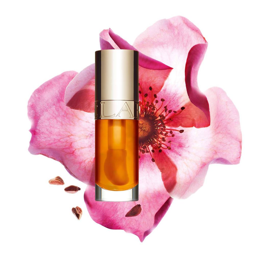 ภาพถ่ายผลิตภัณฑ์ Clarins Lip Comfort Oil สี Honey วางอยู่ข้าง Sweetbriar Rose บนพื้นหลังสีพีชอ่อน