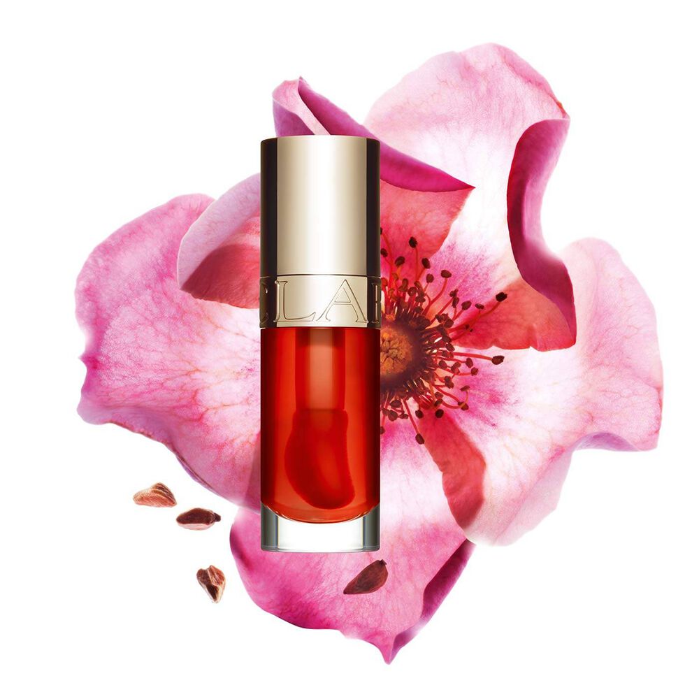 ภาพถ่ายผลิตภัณฑ์ Clarins Lip Comfort Oil Apricot วางอยู่ข้าง Sweetbriar Rose บนพื้นหลังสีพีชอ่อนปนส้ม
