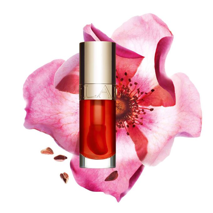 ภาพถ่ายผลิตภัณฑ์ Clarins Lip Comfort Oil Apricot วางอยู่ข้าง Sweetbriar Rose บนพื้นหลังสีพีชอ่อนปนส้ม