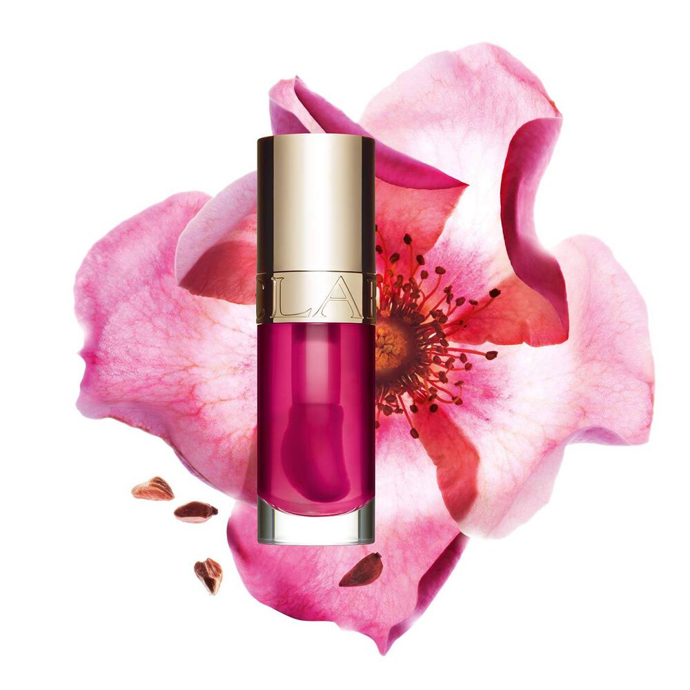 ภาพถ่ายผลิตภัณฑ์ Clarins Lip Comfort Oil สี Raspberry วางอยู่ข้าง Sweetbriar Rose บนพื้นหลังสีพีชอ่อน