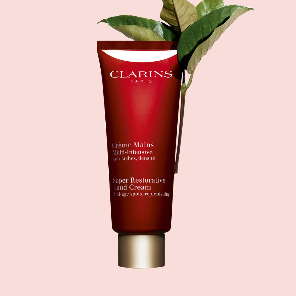 ภาพถ่ายของขวด Clarins Super Restorative Hand Cream ที่วางใกล้ใบฮารุงกานา บนพื้นหลังสีชมพูอ่อน เพื่อเน้นถึงส่วนผสมจากธรรมชาติและคุณสมบัติในการฟื้นบำรุงของผลิตภัณฑ์