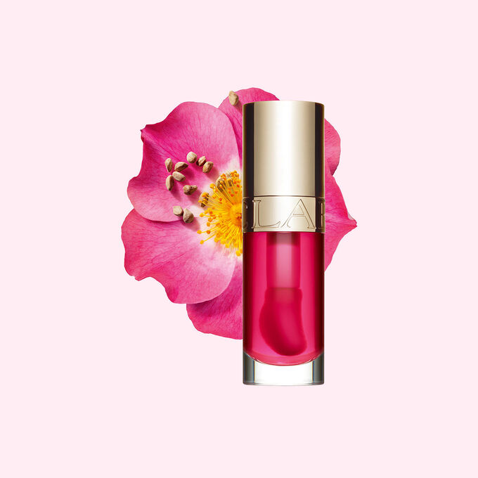 ภาพถ่ายผลิตภัณฑ์ Clarins Lip Comfort Oil สี Pitaya วางอยู่ข้าง Sweetbriar Rose บนพื้นหลังสีชมพูอ่อน