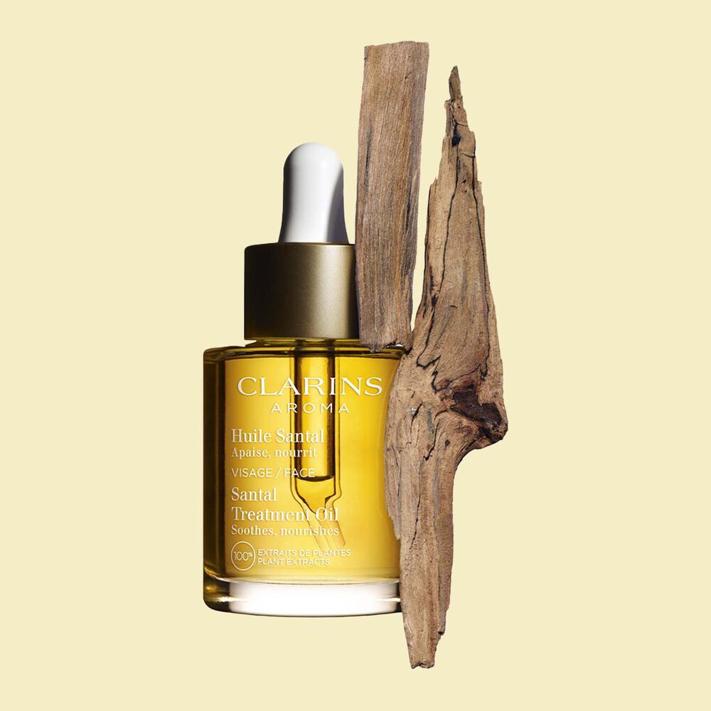 ภาพผลิตภัณฑ์ของขวด Clarins Santal Face Treatment Oil ที่วางใกล้กับเปลือกไม้จันทน์บนพื้นหลังสีเหลืองอ่อน