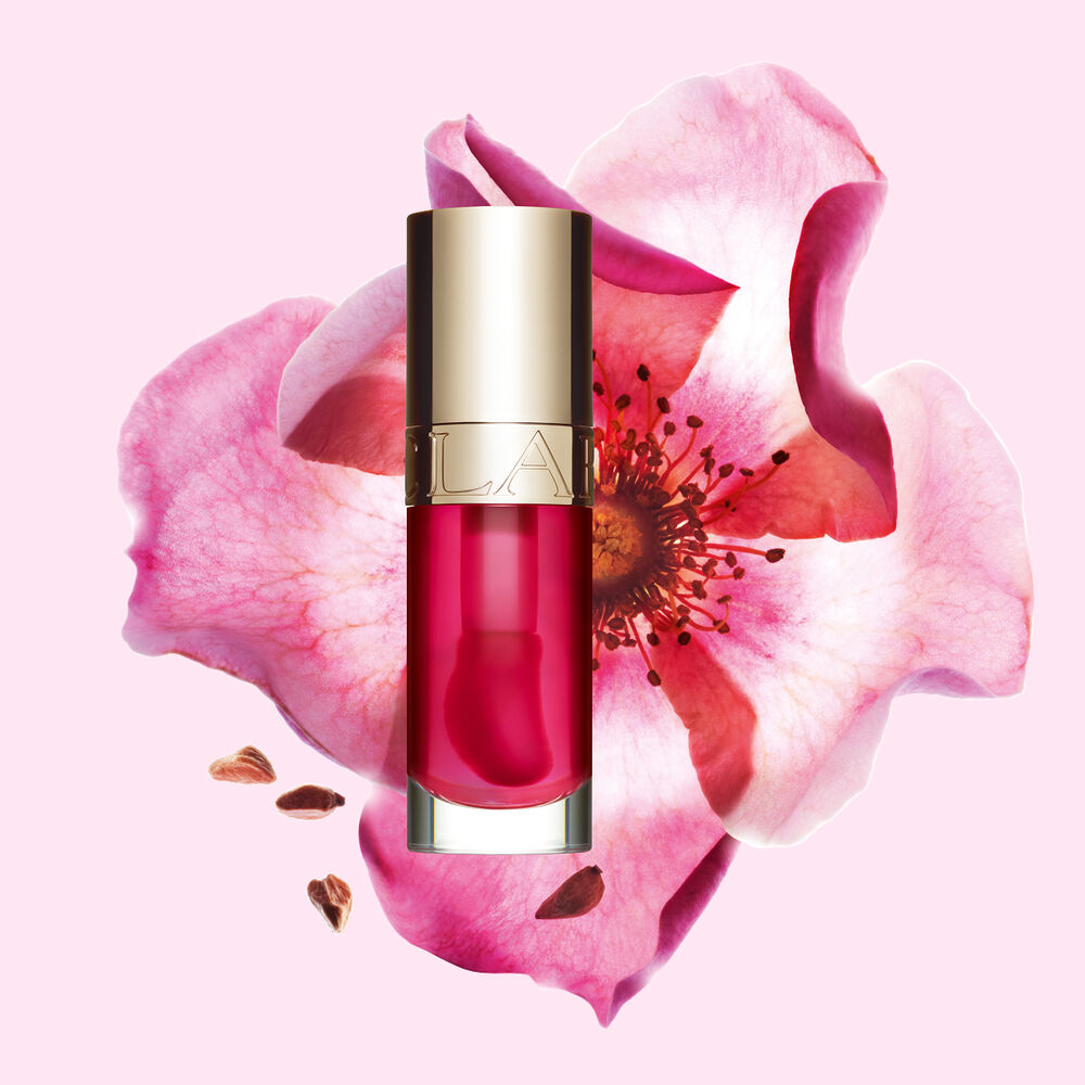 ภาพถ่ายผลิตภัณฑ์ Clarins Lip Comfort Oil สี Pitaya วางอยู่ข้าง Sweetbriar Rose บนพื้นหลังสีชมพูอ่อน