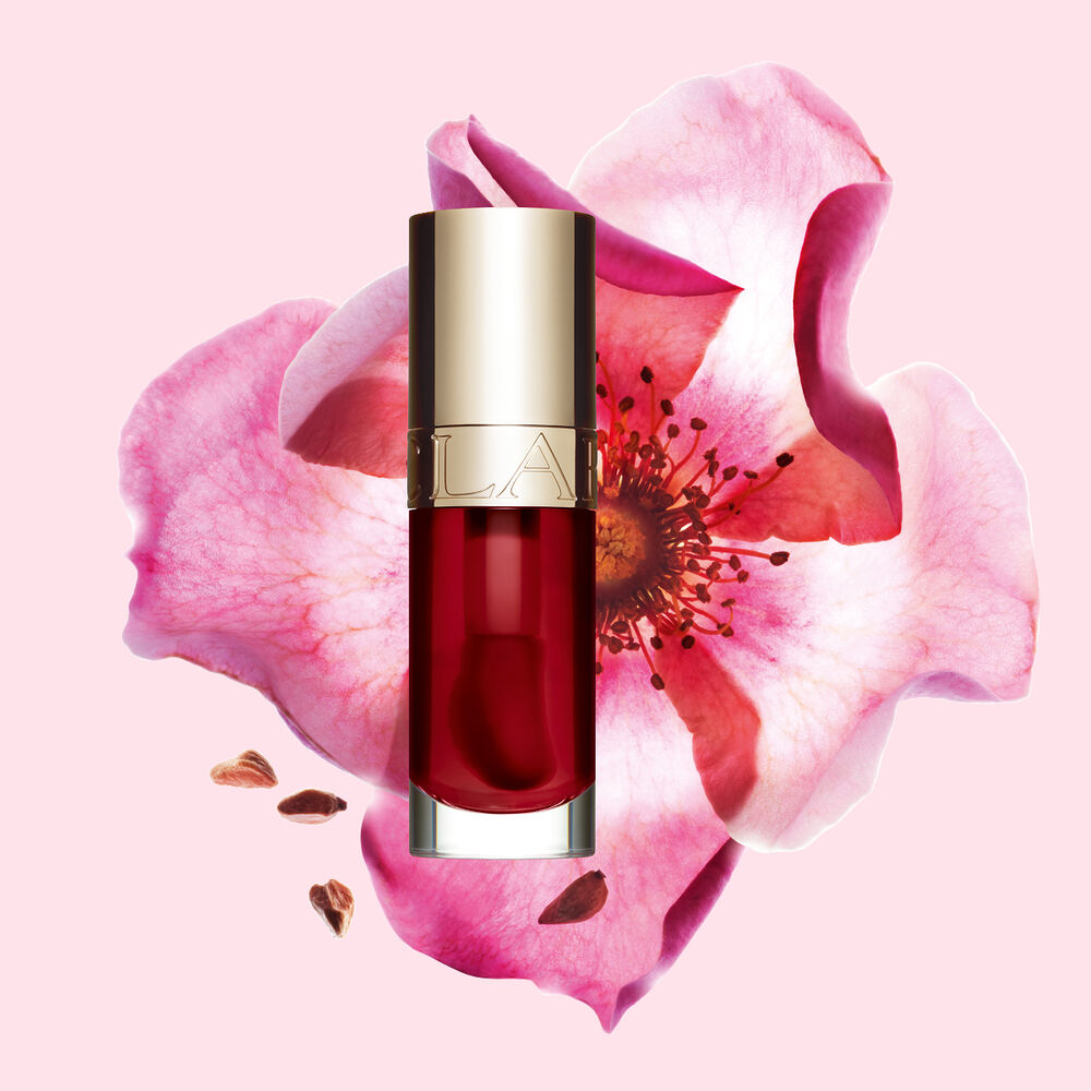 ภาพถ่ายผลิตภัณฑ์ Clarins Lip Comfort Oil สี Cherry วางอยู่ข้าง Sweetbriar Rose บนพื้นหลังสีชมพูอ่อน