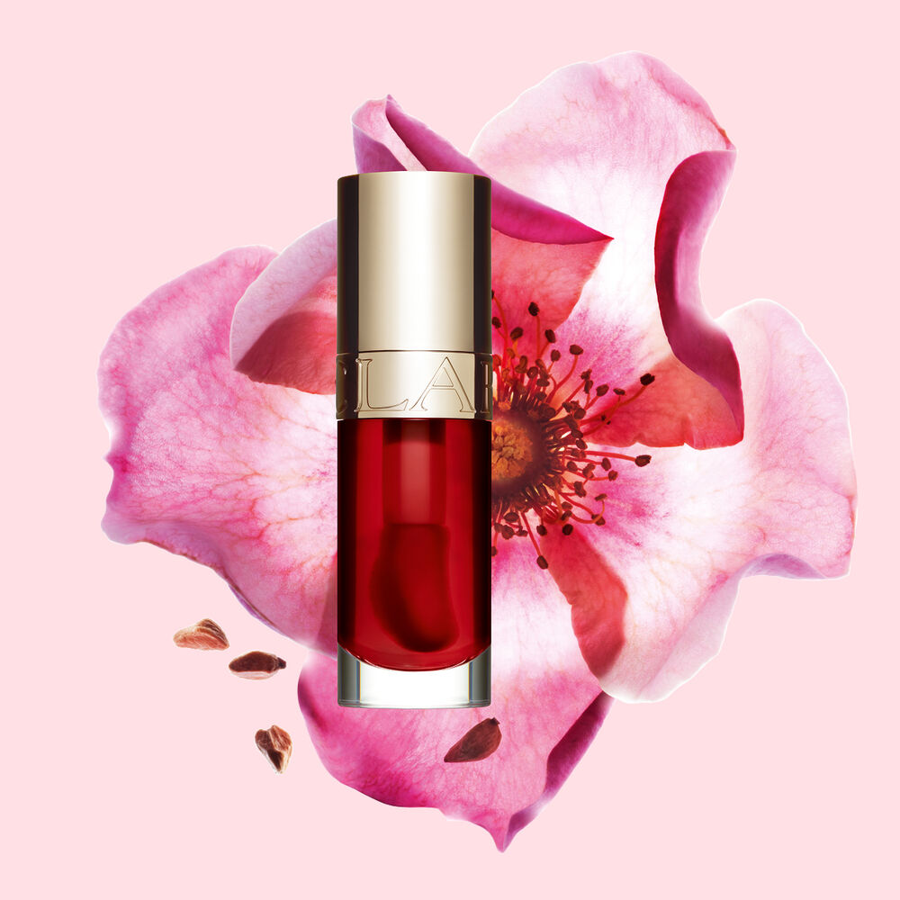 ภาพถ่ายผลิตภัณฑ์ Clarins Lip Comfort Oil Strawberry วางอยู่ข้าง Sweetbriar Rose บนพื้นหลังสีชมพูอ่อน
