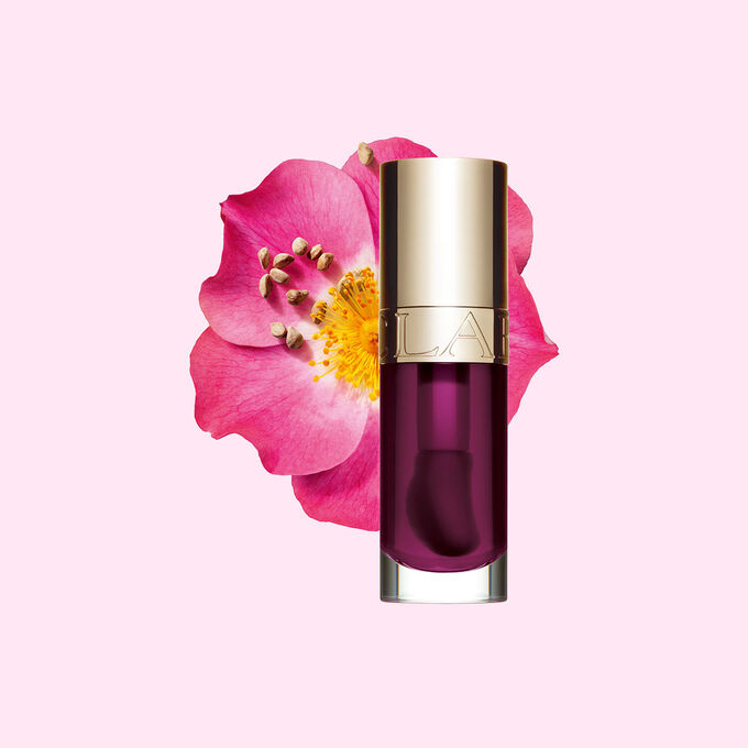 ภาพถ่ายผลิตภัณฑ์ Clarins Lip Comfort Oil Plum วางอยู่ข้าง Sweetbriar Rose บนพื้นหลังสีชมพูอ่อน