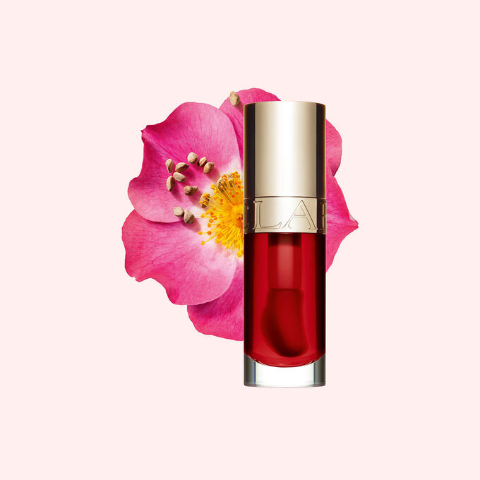 ภาพถ่ายผลิตภัณฑ์ Clarins Lip Comfort Oil Strawberry วางอยู่ข้าง Sweetbriar Rose บนพื้นหลังสีชมพูอ่อน