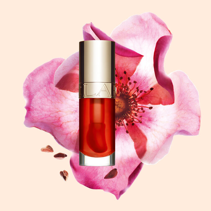 ภาพถ่ายผลิตภัณฑ์ Clarins Lip Comfort Oil Apricot วางอยู่ข้าง Sweetbriar Rose บนพื้นหลังสีพีชอ่อนปนส้ม